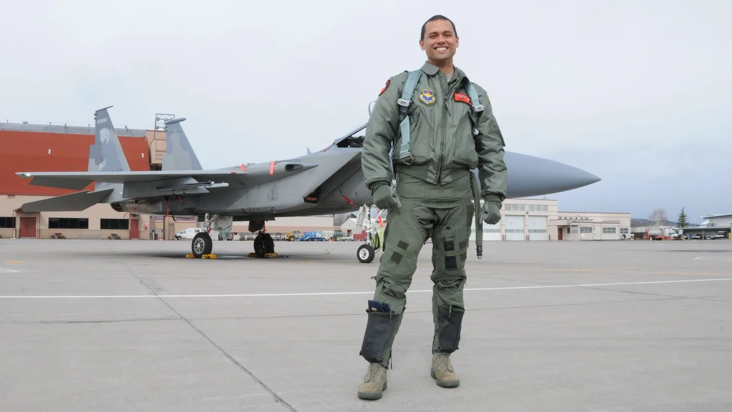 ein Mann in einem Militärpilotenanzug posiert vor einem Düsenflugzeug und Hangars