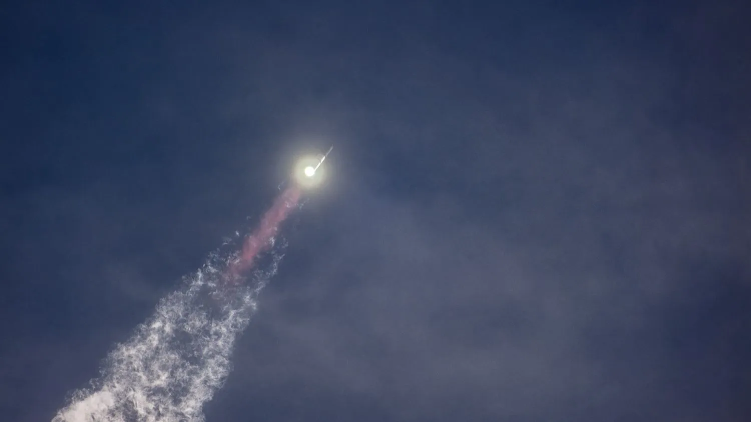 eine große silberne Rakete fliegt durch einen blauen Himmel über einer Flammensäule