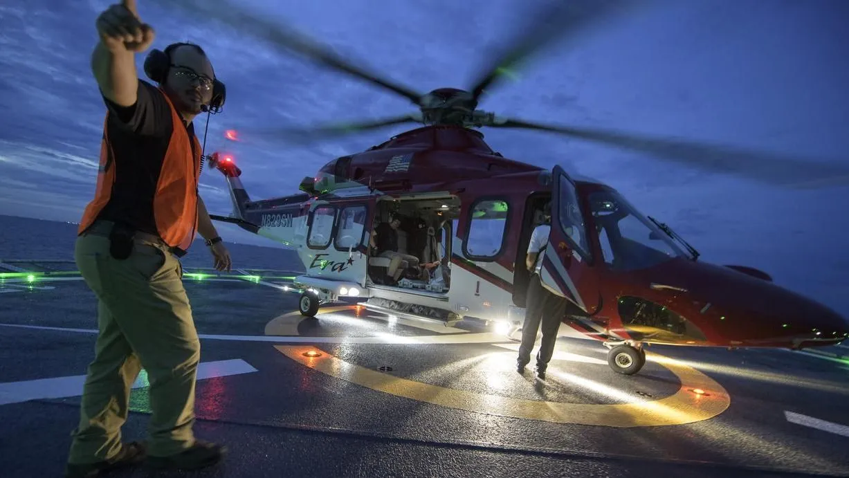 ein Hubschrauber, der sich in der Abenddämmerung auf einem Schiff dreht, mit einer Person davor, die in den Himmel zeigt