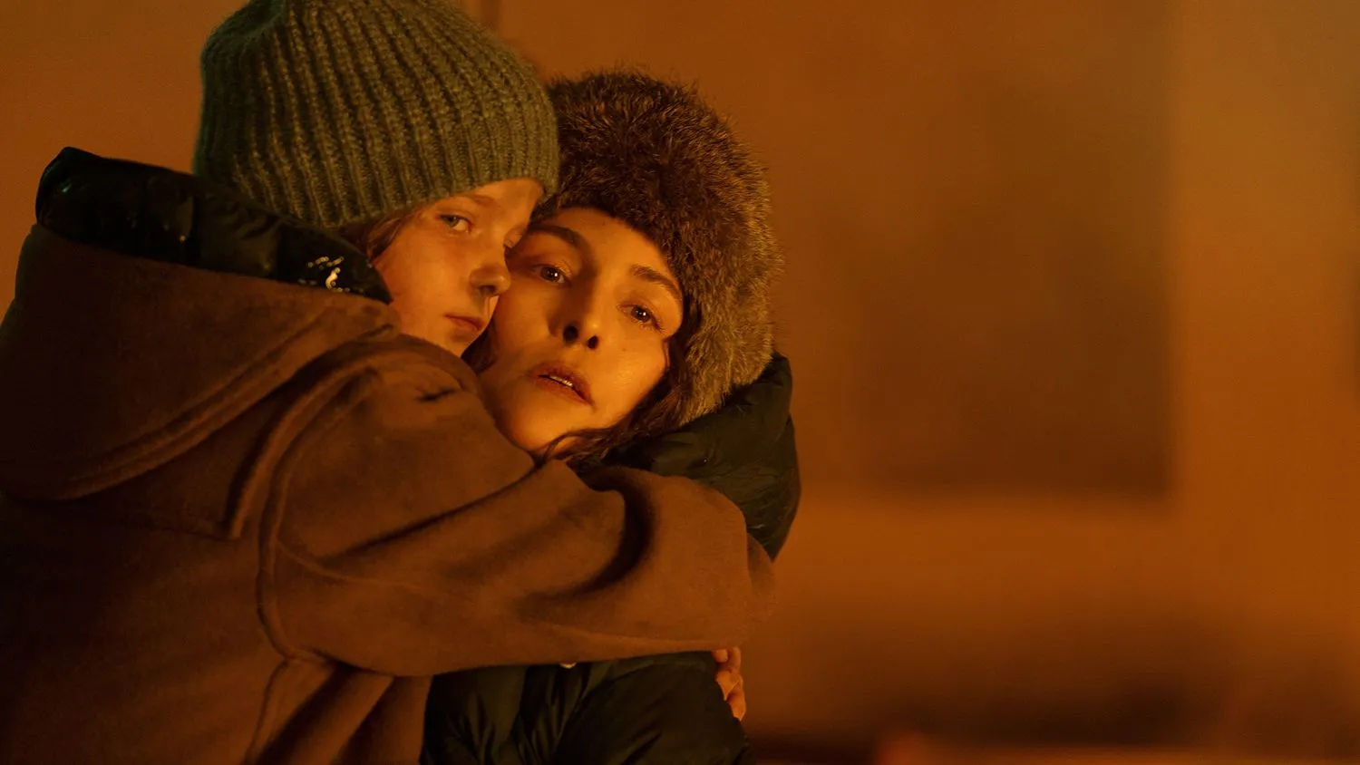 Eine Mutter hebt ihre kleine Tochter hoch und umarmt sie fest. Beide tragen eine warme Wintermütze und eine Jacke. Ihre Gesichter sind mit Ruß bedeckt. Im Hintergrund ist der bernsteinfarbene Schein eines Feuers zu sehen.