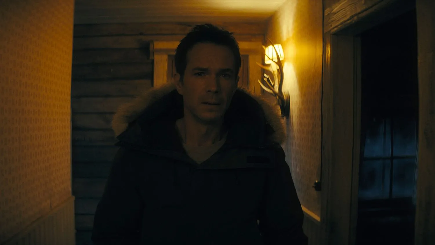 Ein Mann trägt eine dicke Winterjacke, als er nachts eine schwach beleuchtete Holzhütte durchsucht.