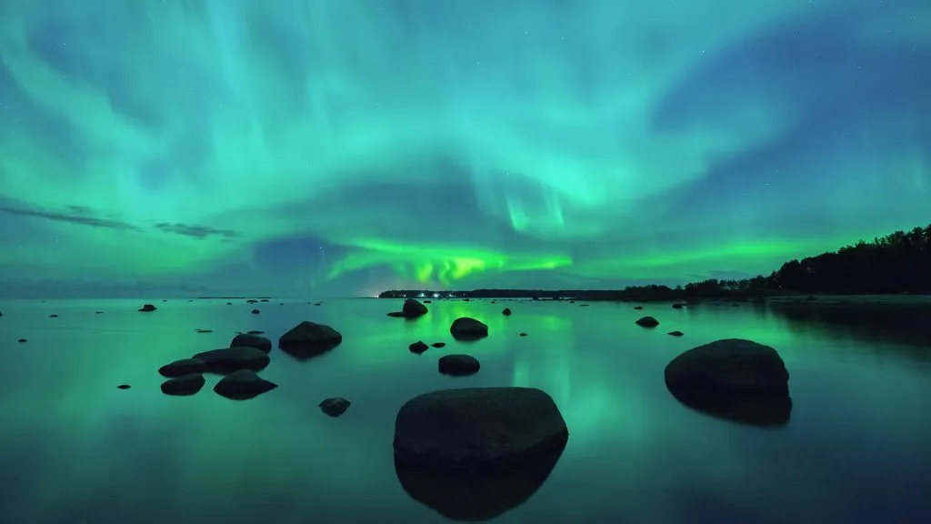 Ein blaugrüner Himmel mit Schlieren von Polarlichtern über einem Gewässer mit felsigen Silhouetten.