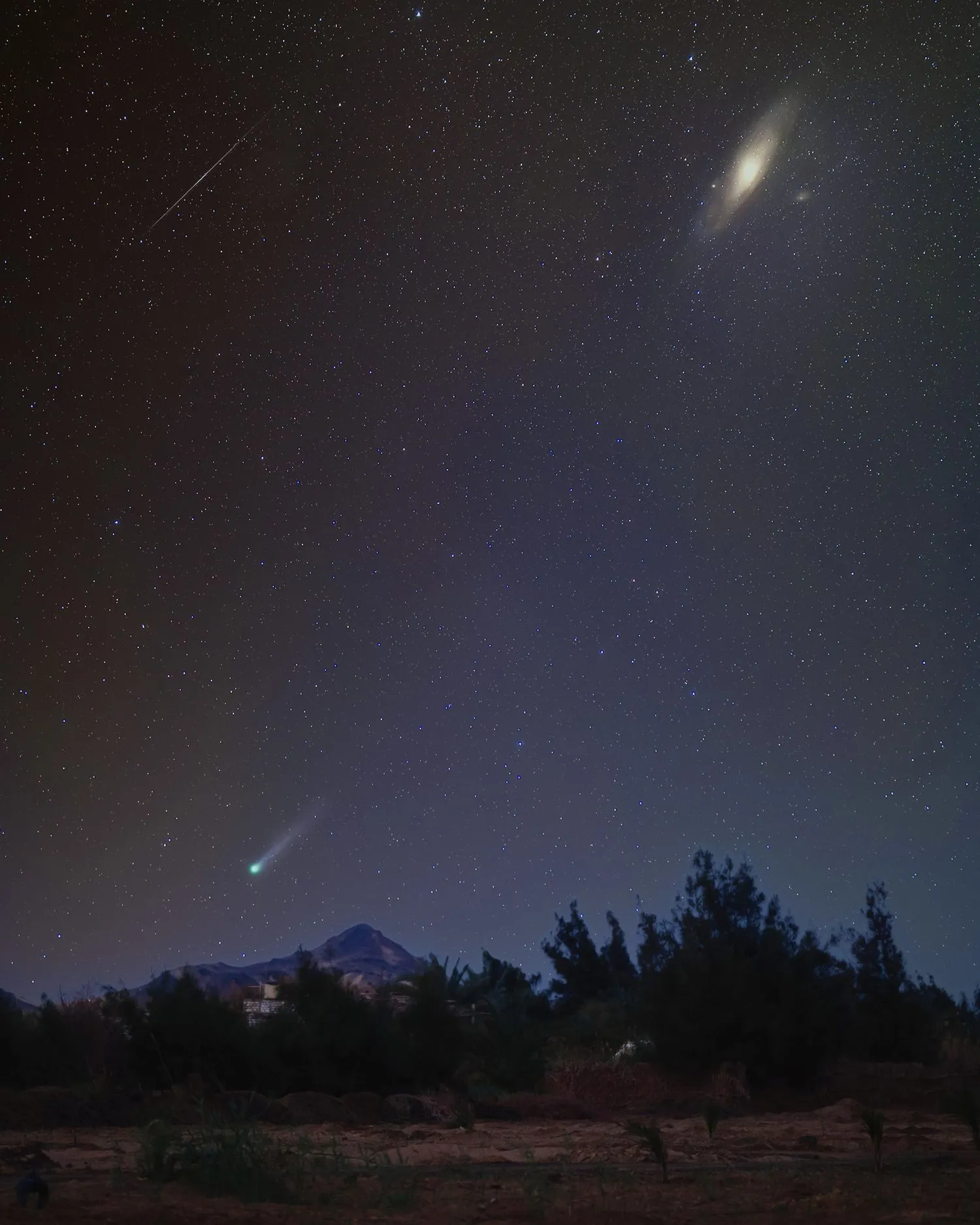 Komet streift über den Himmel und leuchtet grün, mit einer hellen Galaxie in der oberen linken Ecke und einer langen dünnen Meteorspur am Himmel in der oberen linken Ecke.