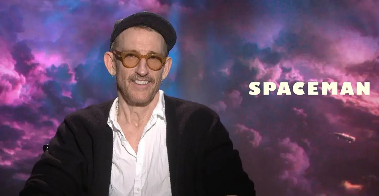 ein Mann mit Hut und Brille sitzt vor einer Grafik eines Raumschiffs, das durch lila Wolken fliegt