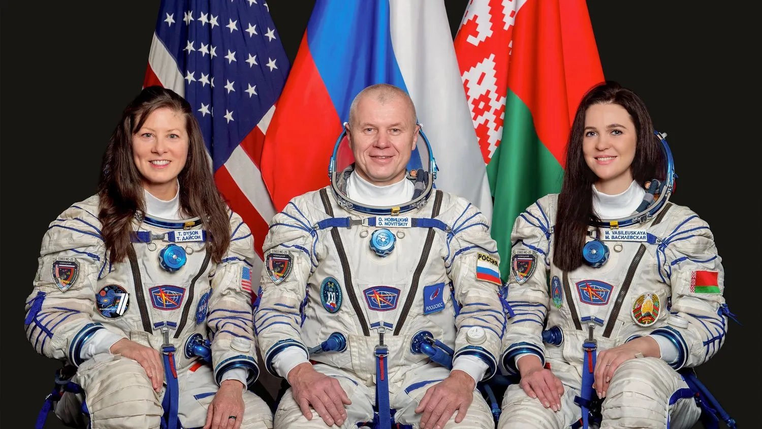 drei lächelnde Astronauten (zwei Frauen und ein Mann) in weißen Raumanzügen sitzen vor ihren Nationalflaggen vor einem dunklen Hintergrund